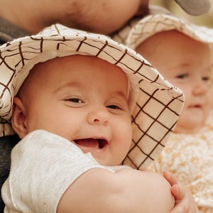 Puede incluir: Primer plano de un padre sosteniendo a dos bebés. Los bebés llevan gorras de sol a juego de cuadros marrones y blancos. El bebé en primer plano sonríe y mira a la cámara.