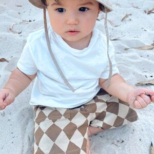 Puede incluir: Un ni&ntilde;o peque&ntilde;o con una camiseta blanca y pantalones cortos a cuadros marrones y blancos se sienta en una playa de arena. El ni&ntilde;o lleva un sombrero de cubo beige con una cuerda marr&oacute;n.