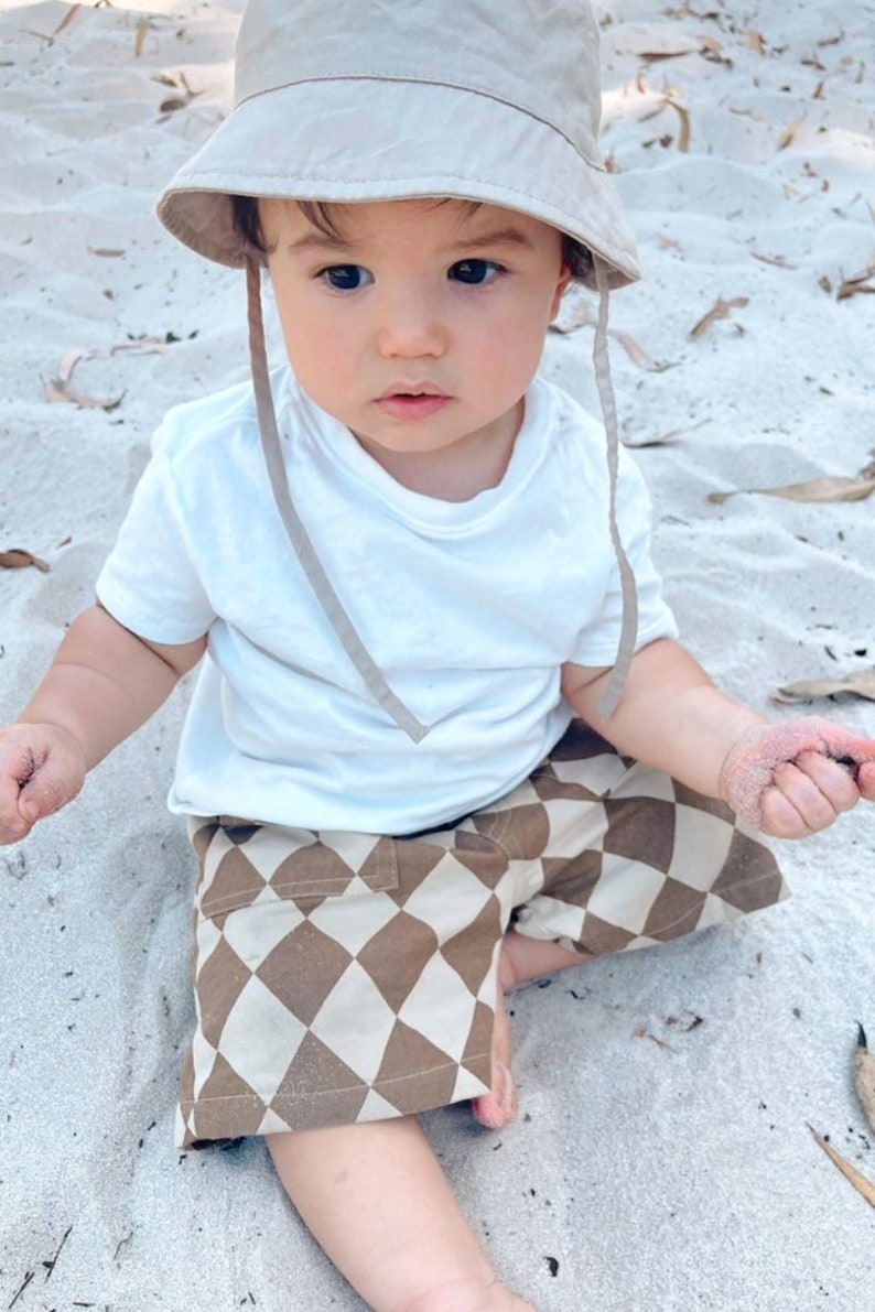 Puede incluir: Un ni&ntilde;o peque&ntilde;o con una camiseta blanca y pantalones cortos a cuadros marrones y blancos se sienta en una playa de arena. El ni&ntilde;o lleva un sombrero de cubo beige con una cuerda marr&oacute;n.