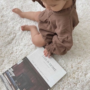 May include: A child in a brown linen dress sits on a white fluffy rug and holds a book titled "This is Home: The Art of Simple Living" by Natalie Walton.