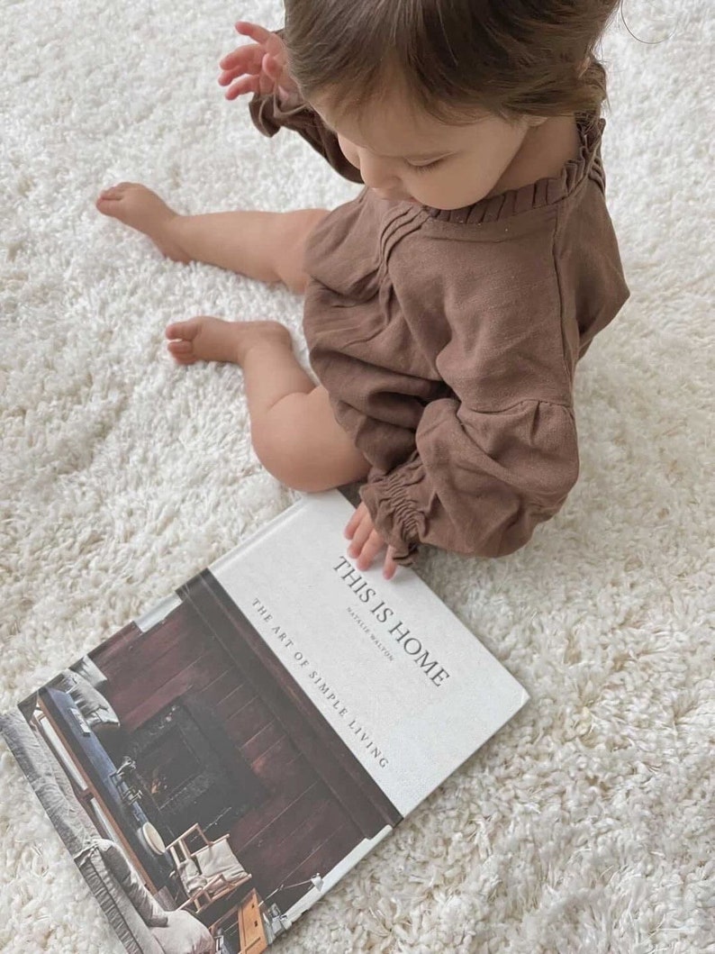 May include: A child in a brown linen dress sits on a white fluffy rug and holds a book titled "This is Home: The Art of Simple Living" by Natalie Walton.