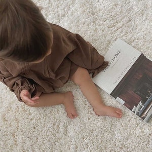 May include: A young child wearing a brown dress sits on a white fluffy rug, looking down at a book titled "This is Home" by  Natalie Walton. The book cover features a photograph of a fireplace with a wooden mantle and a rustic interior.