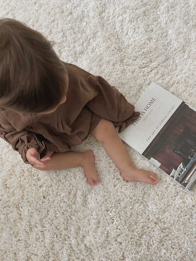 May include: A young child wearing a brown dress sits on a white fluffy rug, looking down at a book titled "This is Home" by  Natalie Walton. The book cover features a photograph of a fireplace with a wooden mantle and a rustic interior.
