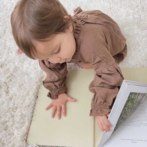 May include: A young child in a brown long-sleeved outfit is sitting on a white fluffy rug and looking at a book with a floral image on the page. The book is open and the child is touching the page with their hand.