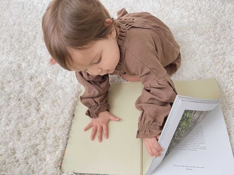 May include: A young child in a brown long-sleeved outfit is sitting on a white fluffy rug and looking at a book with a floral image on the page. The book is open and the child is touching the page with their hand.