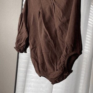 May include: A brown long-sleeved bodysuit with a ruffled neckline and a gathered waist. The bodysuit has a snap closure at the bottom.