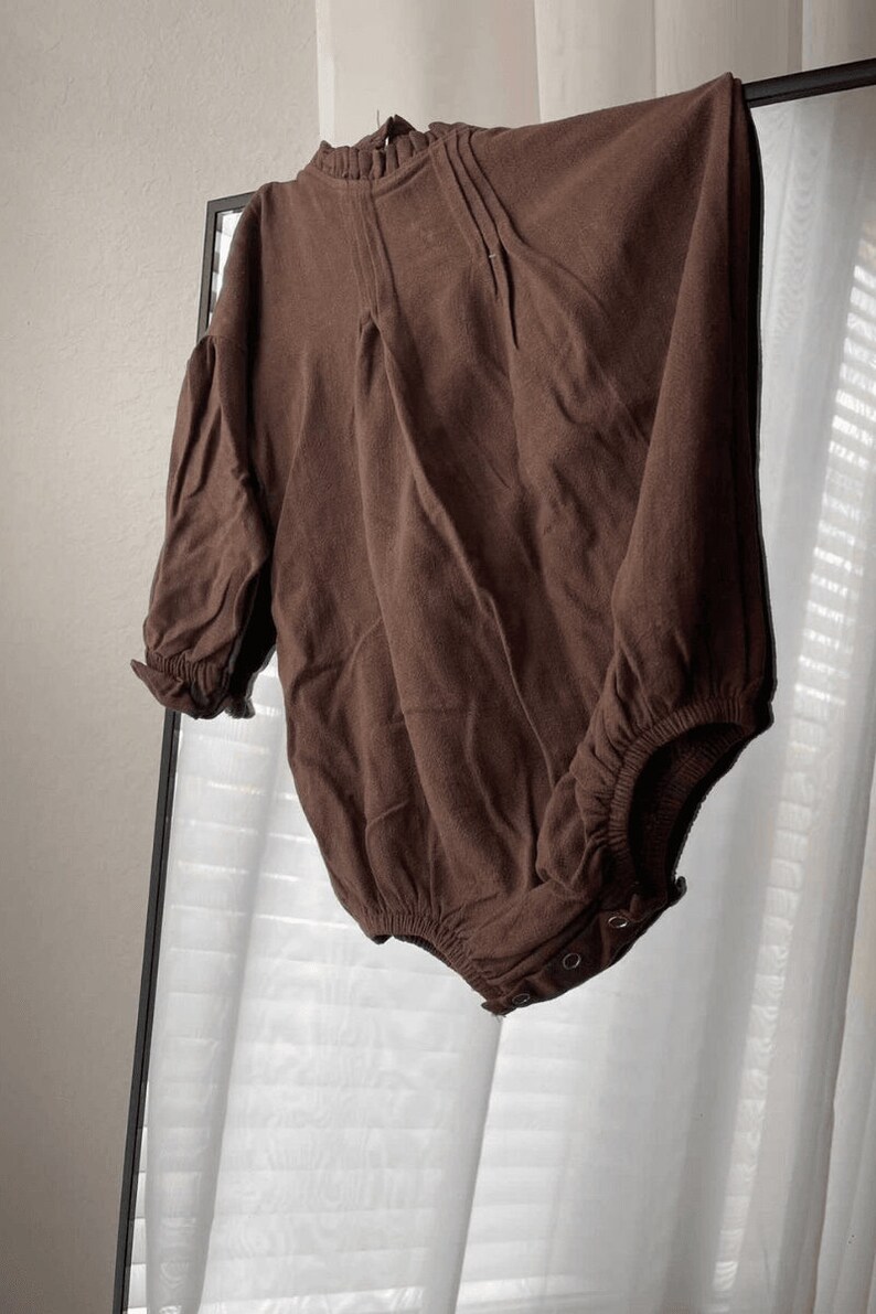 May include: A brown long-sleeved bodysuit with a ruffled neckline and a gathered waist. The bodysuit has a snap closure at the bottom.