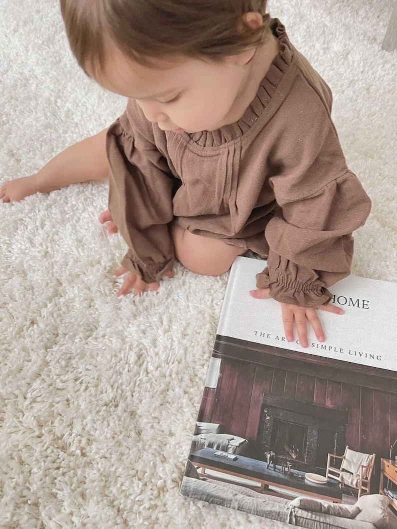 May include: A young child wearing a brown long-sleeved shirt sits on a white fluffy rug and looks at a book titled "The Art of Simple Living: Home".