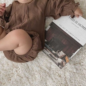 May include: A baby wearing a brown linen romper is holding a book titled "This is Home: The Art of Simple Living" by Natalie Walton.