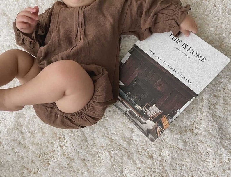May include: A baby wearing a brown linen romper is holding a book titled "This is Home: The Art of Simple Living" by Natalie Walton.