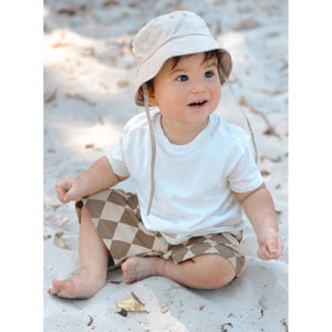 Puede incluir: Un beb&eacute; con una camiseta blanca, pantalones cortos a cuadros marrones y beige y un sombrero de cubo beige se sienta en una playa de arena.