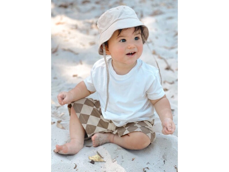 Puede incluir: Un beb&eacute; con una camiseta blanca, pantalones cortos a cuadros marrones y beige y un sombrero de cubo beige se sienta en una playa de arena.