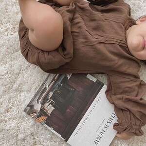 May include: A brown baby onesie with long sleeves and a ruffled neckline. The onesie is lying on a white fluffy rug with a book titled "This Is Home" by Melissa Hillen, "The Art of Simple Living" on the cover.