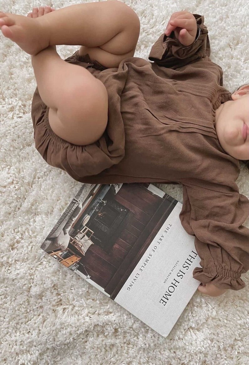 May include: A brown baby onesie with long sleeves and a ruffled neckline. The onesie is lying on a white fluffy rug with a book titled "This Is Home" by Melissa Hillen, "The Art of Simple Living" on the cover.