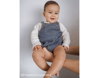 May include: A baby wearing a white and blue knitted sweater and denim overalls is being held up by an adult's hands. The baby is smiling and looking at the camera. The background is a light gray wall. @threelilttlepeople_