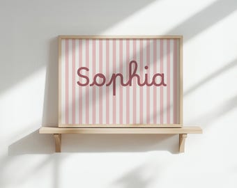May include: A framed print featuring the name "Sophia" in a cursive font, set against a backdrop of vertical pink and white stripes. The wooden frame sits on a light-colored shelf, creating a simple, elegant display.