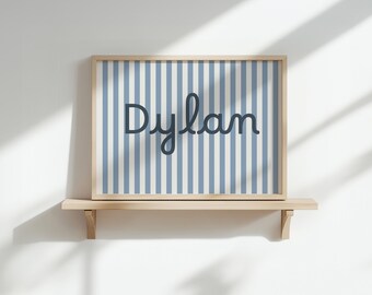 May include: A framed print with the name "Dylan" in dark blue script. The background has blue and white vertical stripes. The frame is light wood, and the print is displayed on a wooden shelf. The overall design is simple.