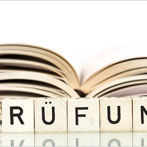 Könnte beinhalten: Holzbuchstaben, die das deutsche Wort „PRÜFUNG“ (Prüfung oder Test) bilden, sind vor einem offenen Buch angeordnet. Die Blöcke sind hellfarbig mit schwarzen Buchstaben. Der Hintergrund ist ein verschwommenes Bild der Buchseiten.