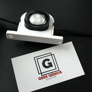 May include: A white, rectangular device with a black, circular lens. The device is connected to a black wire and sits above a business card with the text "GEEK GENIUS COLLECTIVE" and a stylized "G" logo.