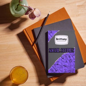 May include: A composition of stationery items on a wooden surface. A notebook with the name "Brittany" and the words "Social Studies" is visible. A pencil, a glass of orange liquid, and a vase with purple flowers are also present.