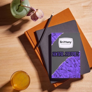 May include: A composition of stationery items on a wooden surface. A notebook with the name "Brittany" and the words "Social Studies" is visible. A pencil, a glass of orange liquid, and a vase with purple flowers are also present.