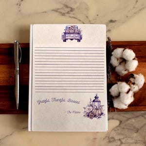 May include: A lined journal with a white cover featuring a purple illustration of a truck carrying flowers. The journal includes the text "Grateful, Thankful, Blessed" and "- The Millers". A silver pen rests beside the journal.