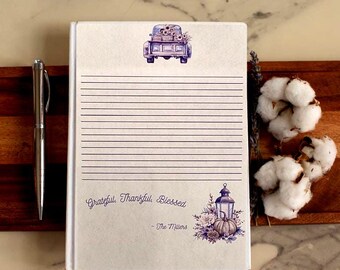 May include: A lined journal with a white cover featuring a purple illustration of a truck carrying flowers. The journal includes the text "Grateful, Thankful, Blessed" and "- The Millers". A silver pen rests beside the journal.