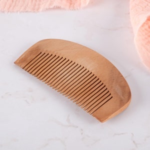 May include: A wooden comb with a curved shape and fine teeth. The comb is made of light brown wood and is lying on a white marble surface.