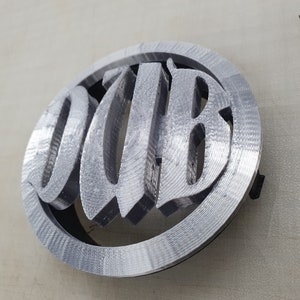Puede incluir: Un monograma de metal plateado con las letras "OUB" en un diseño circular.
