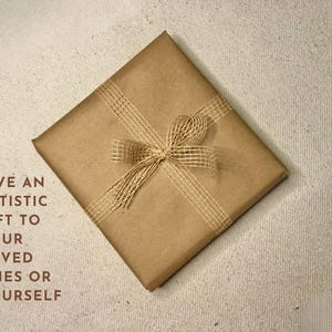K&ouml;nnte beinhalten: Eine braune Papiert&uuml;te mit einer Schleife aus Naturbast. Auf der T&uuml;te steht der Text "GIVE AN ARTISTIC GIFT TO YOUR LOVED ONES OR YOURSELF".