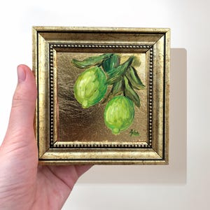 May include: A framed painting of two green limes on a branch with green leaves. The painting is on a gold background.