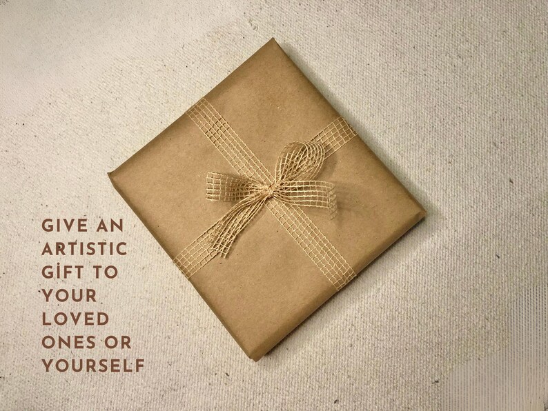 Op de afbeelding: Een vierkant cadeau verpakt in bruin papier, versierd met een geweven lint en strik. De tekst luidt: "GEEF EEN ARTISTIEK CADEAU AAN UW GELIEFDE OF AAN UZELF."