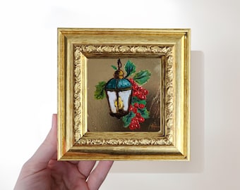 May include: A framed painting of a teal and white lantern with a lit candle, red berries, and green leaves. The painting is in a gold ornate frame. The artwork is held by a hand.