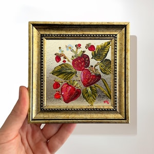 Op de afbeelding: Een kleine, vierkante schildering van aardbeien in een gouden lijst. Het kunstwerk toont levendige rode aardbeien met groene bladeren en kleine witte bloemen. De lijst heeft een gedetailleerd, sierlijk ontwerp.