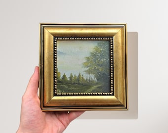 May include: A small framed painting of a landscape with trees and a cloudy sky. The frame is gold with a beaded border.