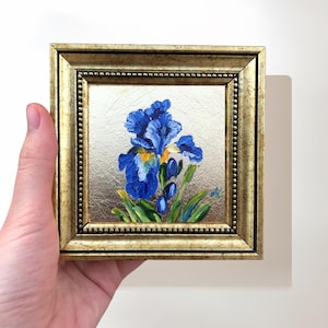 May include: A small, square framed painting of a blue iris flower. The painting has a gold-colored frame with a beaded edge. The iris is painted with shades of blue, yellow, and green against a gold background.