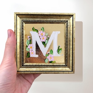 May include: Square framed artwork featuring the letter "M" in white, adorned with pink flowers and green leaves. The artwork is set against a gold background and framed in a gold-colored frame. The piece is held by a hand.