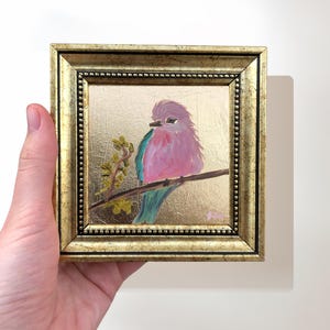 May include: A small painting of a pink bird with a blue tail perched on a branch. The painting is framed in a gold frame.