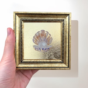 May include: Square framed artwork featuring a gold background and a painted seashell with a pearl. The frame is gold with a beaded border. The seashell is painted in shades of orange, white, and blue.