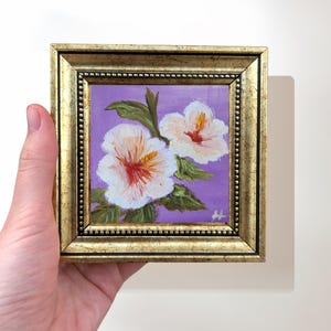 May include: Small framed painting of two white hibiscus flowers with red centers and green leaves against a purple background. The painting is in a gold-colored frame. The artwork is held by a hand.