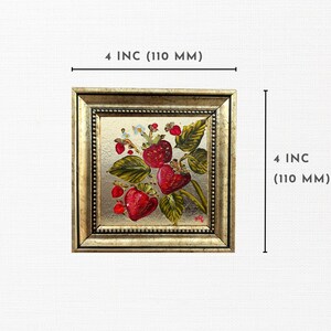 Op de afbeelding: Vierkant ingelijst kunstwerk met aardbeien. De goudkleurige lijst omringt een schilderij van rijpe rode aardbeien met groene bladeren en witte bloemen. Het kunstwerk meet 10 cm per zijde.