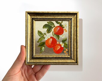May include: Small framed painting featuring three vibrant orange fruits with green leaves and white blossoms. The artwork is set against a gold background and framed in a gold-colored ornate frame. A decorative piece for home decor.