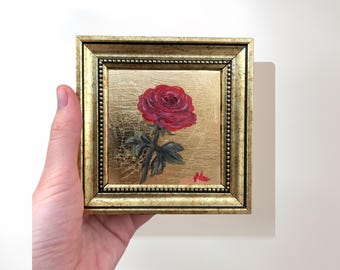 May include: A framed painting of a single red rose with green leaves on a gold background.