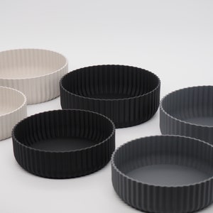 Könnte beinhalten: Sechs runde Schalen mit geripptem Design in verschiedenen neutralen Farben. Die Schalen sind in den Farbtönen Off-White, Schwarz und Grau gehalten. Die Schalen haben ein schlichtes, modernes Design und sind vielseitig einsetzbar.