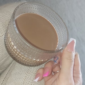 May include: A glass mug with a textured surface filled with brown coffee. The mug is being held by a hand with white and pink nail polish.
