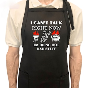 May include: Black apron with white text and graphics. The text reads "I CAN'T TALK RIGHT NOW I'M DOING HOT DAD STUFF." Graphics include a grill, skewers, and a barbecue. The apron has a neck strap and waist ties.