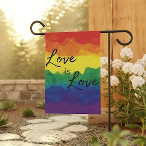 May include: A garden flag featuring a rainbow design with the words "Love is Love" in black script. The flag is displayed on a black metal stand in a garden, surrounded by greenery and white flowers.