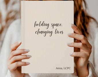 May include: A cream-colored journal with the words "holding space, changing lives" in black script. The name "Anna, LCPC" is printed at the bottom. The journal is held by a person wearing a white sweater.
