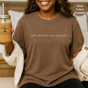 May include: A brown t-shirt with the phrase "radicalized by basic empathy" printed in a light color. The shirt is a solid color and has short sleeves. The image also shows a mason jar with a straw and a drink inside.