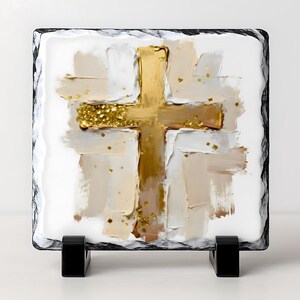May include: Square decorative art piece featuring a gold cross design with gold glitter accents. The cross is set against a backdrop of white and beige brushstrokes, framed by a black border. The art piece is displayed on a black stand.